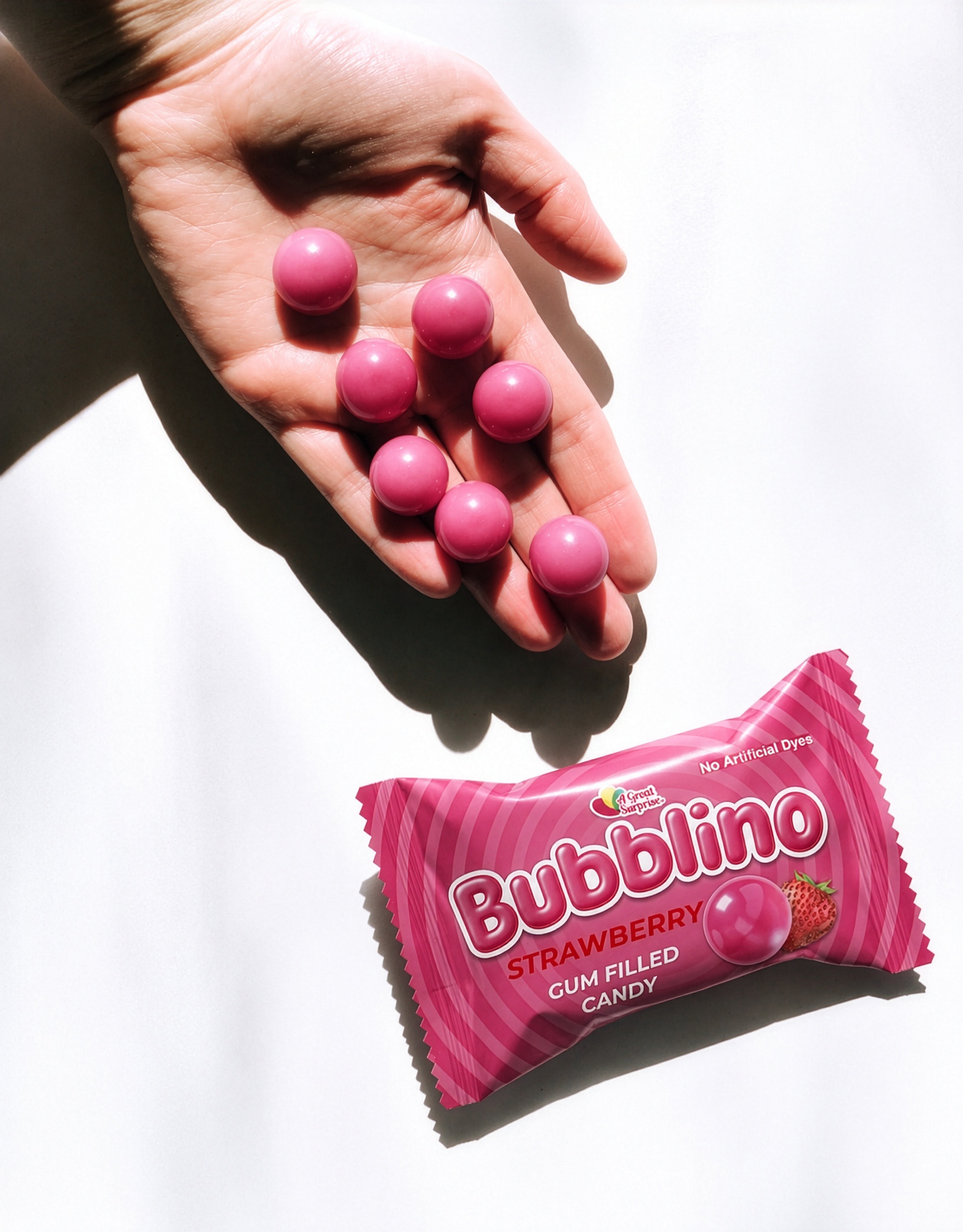A hand releasing pink gumballs above a strawberry wrapper in hard sunlight